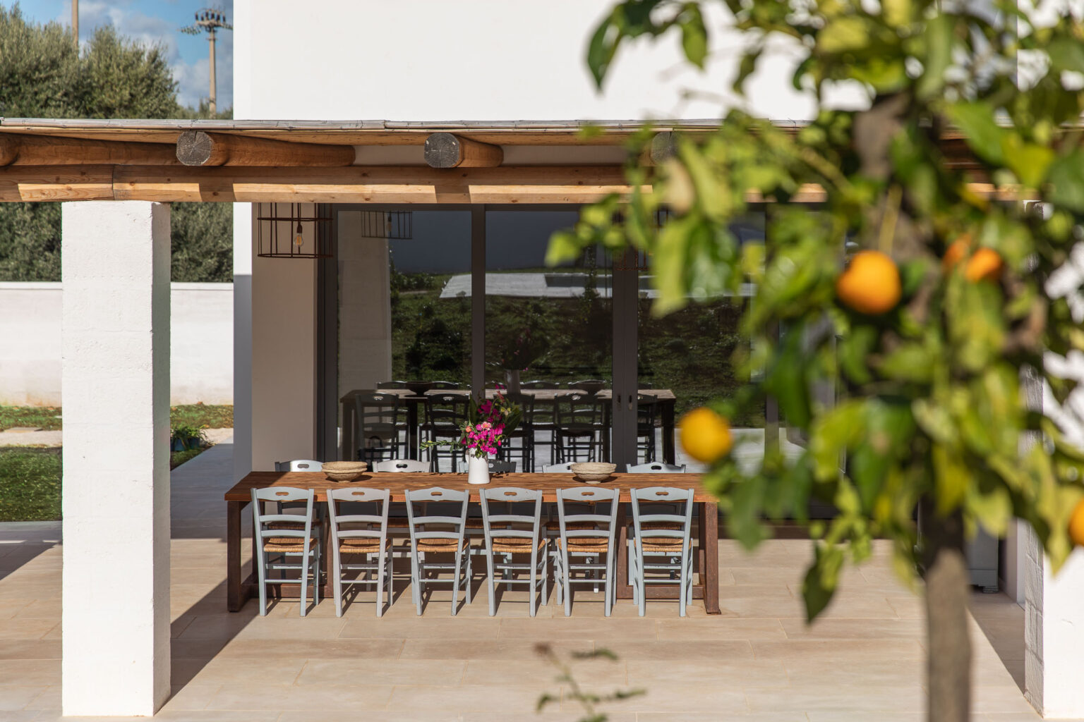 Outdoor dining area with a long wooden table and metal chairs, framed by a modern architectural design and a citrus tree bearing oranges, set in a serene garden environment. | Only on SopranoVillas