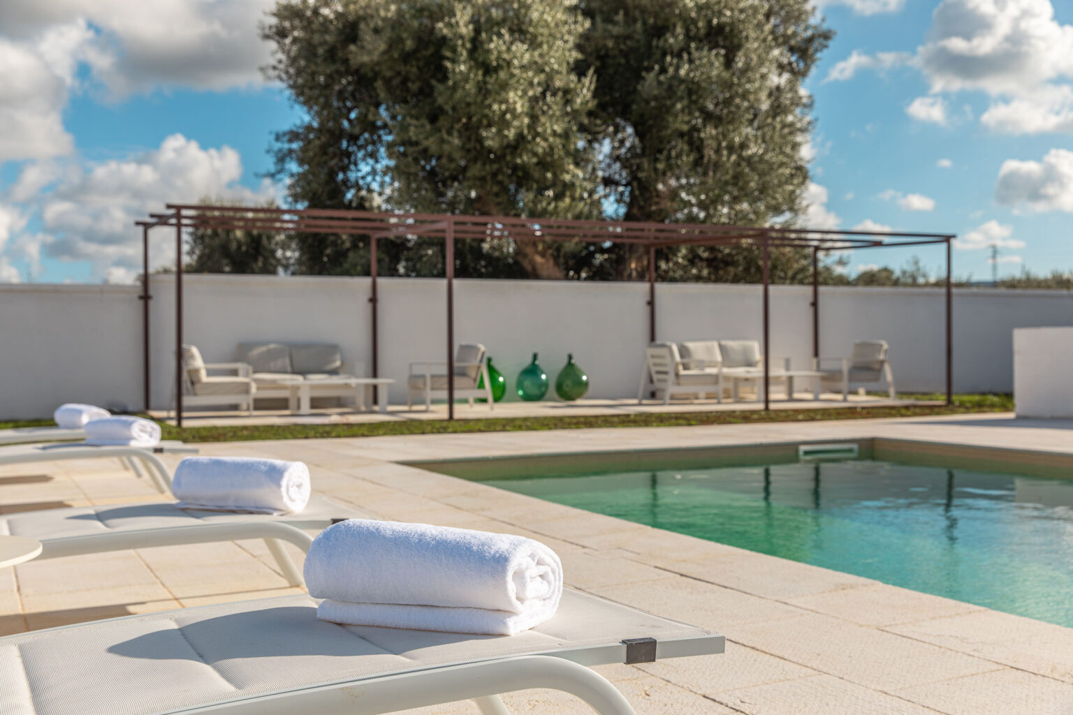 Modern outdoor pool area featuring lounge chairs with rolled towels, a shaded seating area, and decorative green glass vases, set against a backdrop of trees and a blue sky. | Only on SopranoVillas