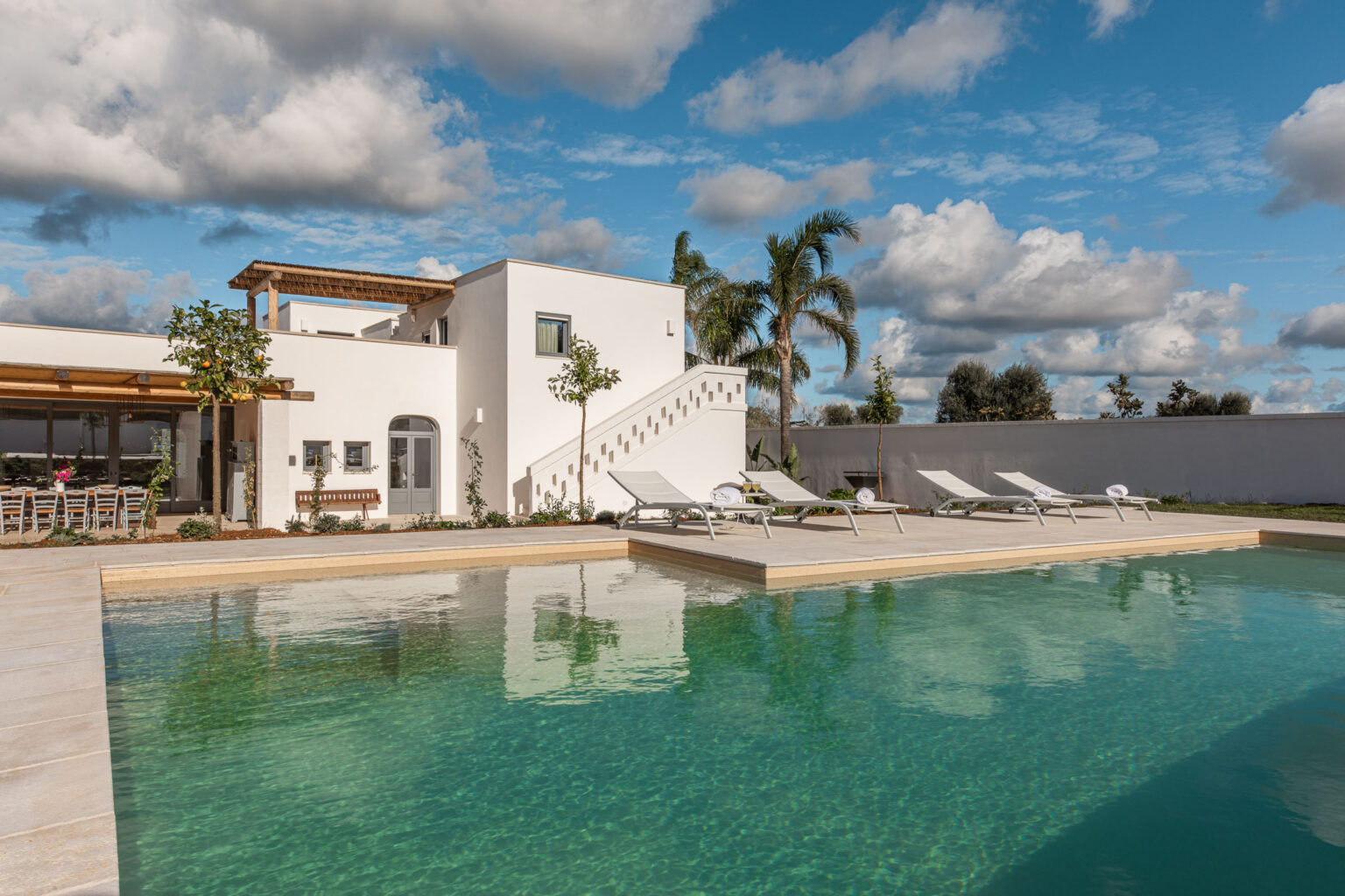 Modern luxury villa with a swimming pool, surrounded by palm trees and a spacious patio, under a blue sky with fluffy clouds. Ideal for relaxation and outdoor gatherings. | Only on SopranoVillas