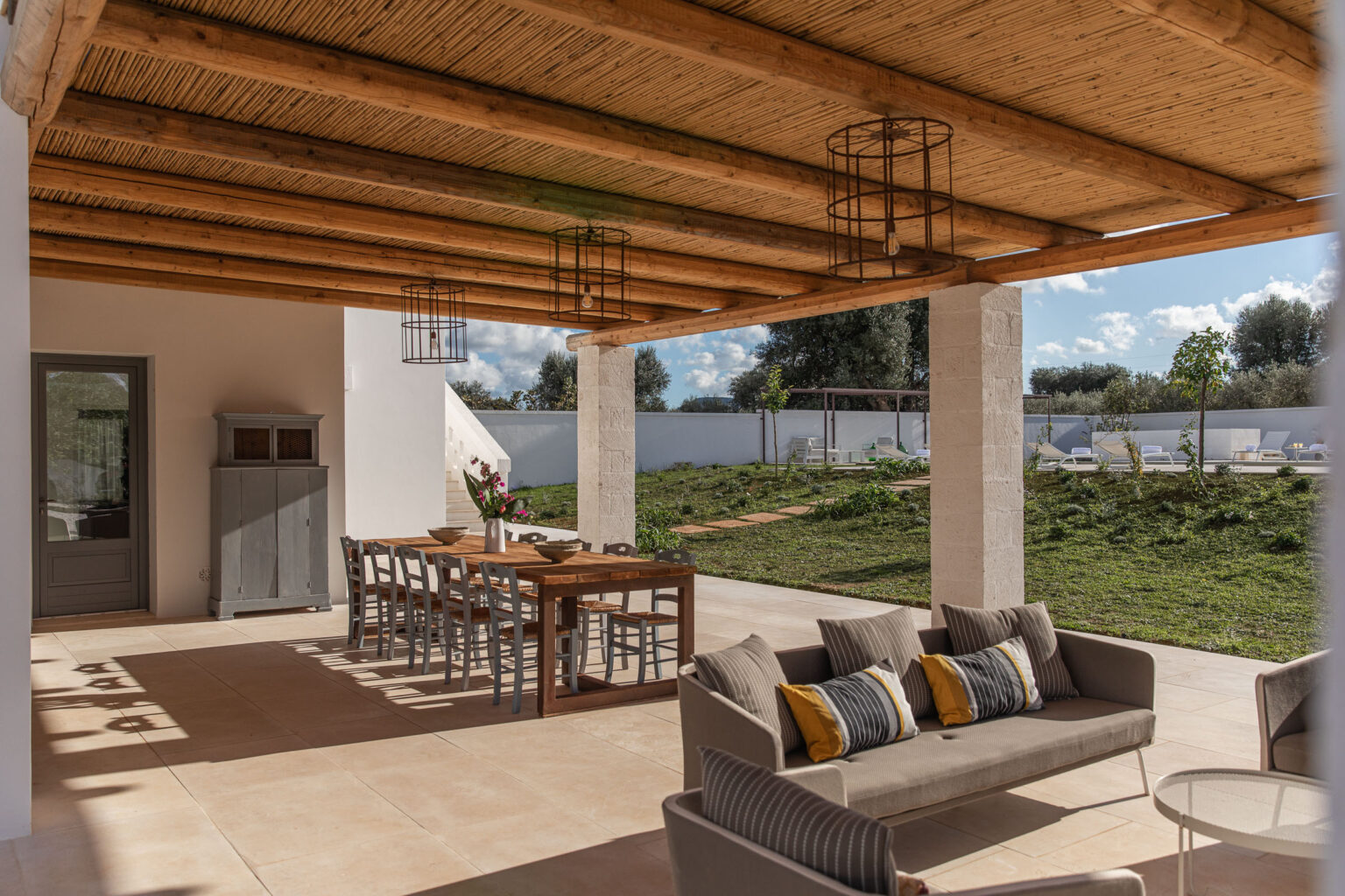 Outdoor patio featuring a cozy seating area with a grey sofa adorned with colorful cushions, complemented by a large wooden dining table. The space is covered with a rustic thatched roof and has a serene green landscape in the background, perfect for entertaining or relaxing. | Only on SopranoVillas