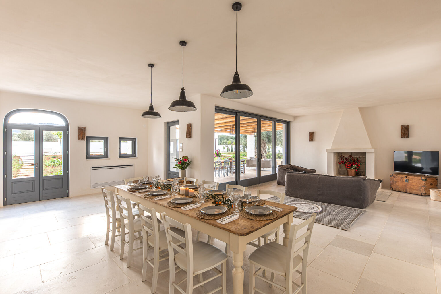 Modern dining room featuring a long wooden table set for a meal, surrounded by white chairs. The space includes large windows with views of the outdoor area, a cozy seating area with a gray sofa, and a stylish fireplace. Industrial-style pendant lights hang from the ceiling, enhancing the bright and airy atmosphere. | Only on SopranoVillas