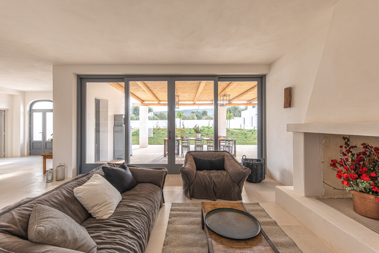 Cozy living room with a plush sofa and armchairs, featuring a modern fireplace and large glass doors that open to a spacious outdoor dining area surrounded by greenery. Natural light illuminates the neutral decor and comfortable furnishings, creating a warm and inviting atmosphere. | Only on SopranoVillas