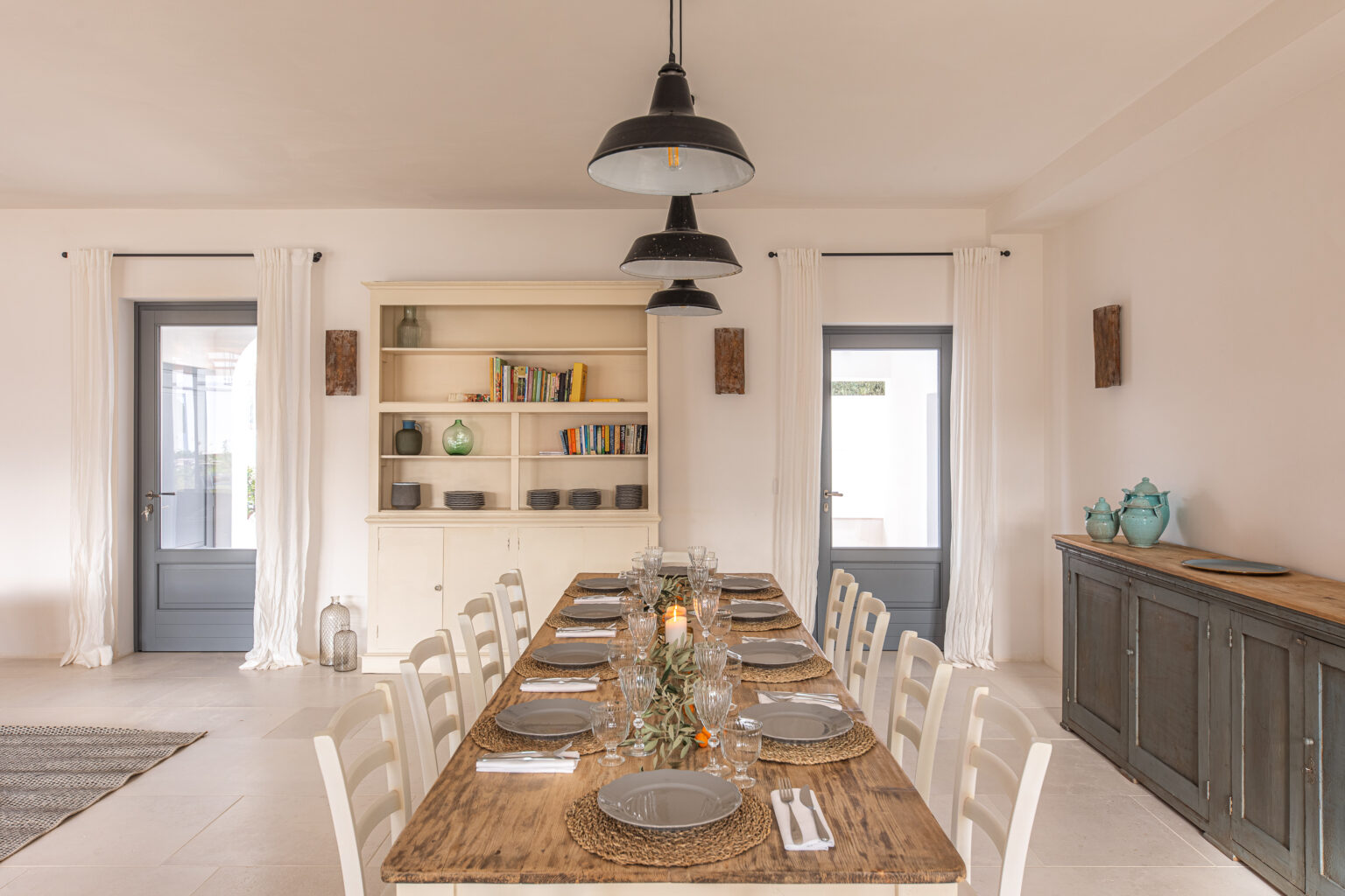 Modern dining room featuring a long wooden table set for a meal with grey plates, glassware, and decorative elements. The space includes white curtains, a light-colored sideboard with turquoise dishes, and a bookshelf with colorful books. Industrial-style pendant lights hang from the ceiling, enhancing the contemporary aesthetic. | Only on SopranoVillas