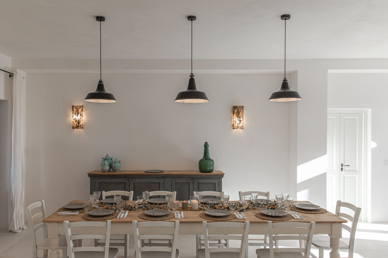 Modern dining room featuring a long wooden table set for a meal, surrounded by white chairs. Stylish black pendant lights and wall sconces illuminate the space, complemented by a gray sideboard and decorative items. | Only on SopranoVillas