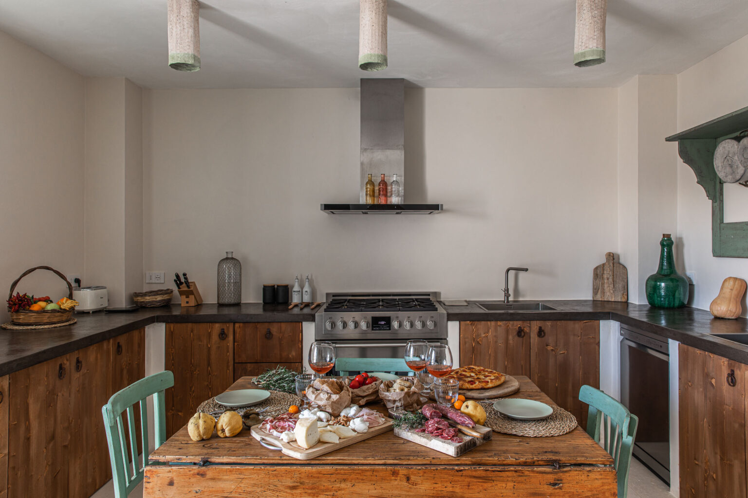 Modern kitchen with rustic wooden cabinetry and a large central table set for a meal, featuring charcuterie, cheeses, fresh fruits, and wine glasses. Stainless steel appliances and decorative elements like glass bottles and wooden cutting boards enhance the inviting atmosphere. | Only on SopranoVillas