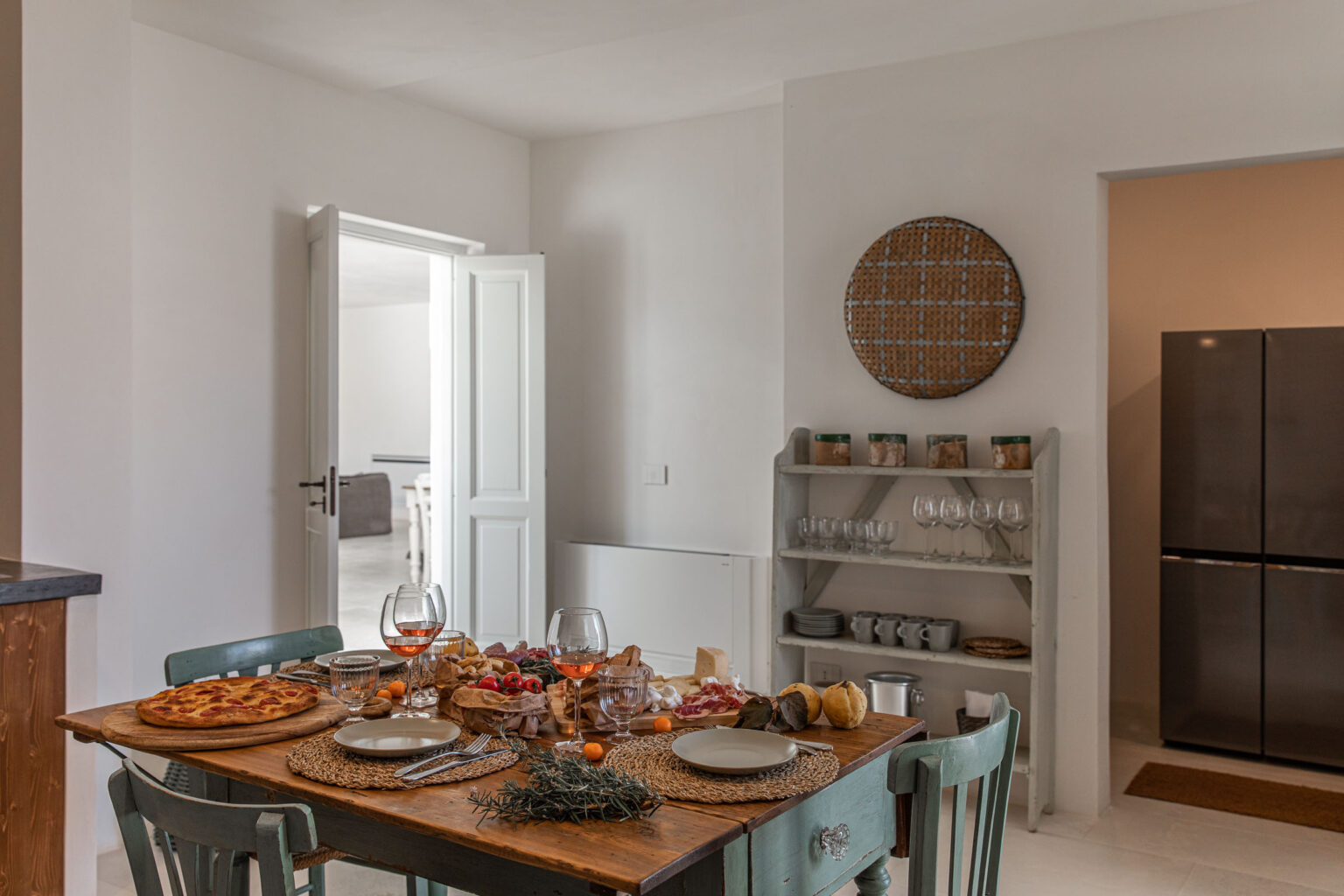 Cozy dining room featuring a wooden table set for a meal, surrounded by teal chairs, with a decorative wall shelf and a modern refrigerator in the background. Natural light fills the space, creating a warm and inviting atmosphere. | Only on SopranoVillas