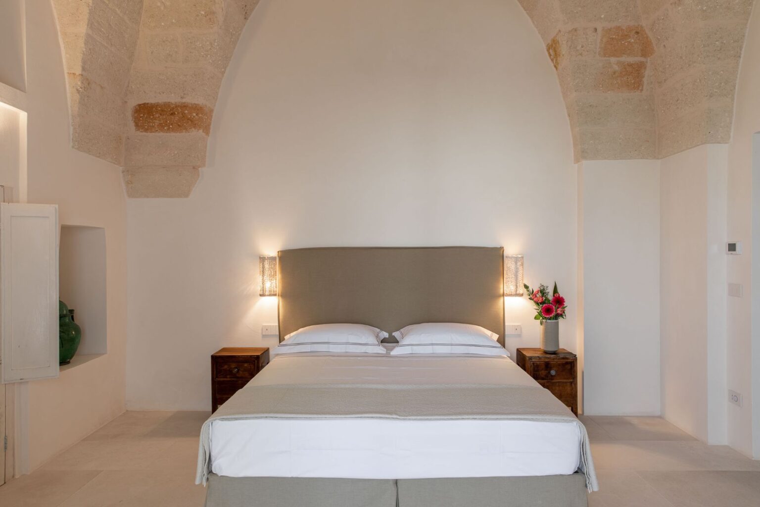 Modern bedroom interior featuring a cozy bed with light-colored linens, soft wall lighting, and natural stone architecture. Decorative flowers on a nightstand add warmth to the serene atmosphere. | Only on SopranoVillas