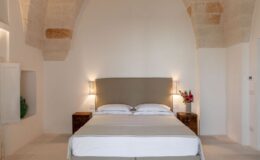 Modern bedroom interior featuring a cozy bed with light-colored linens, soft wall lighting, and natural stone architecture. Decorative flowers on a nightstand add warmth to the serene atmosphere.