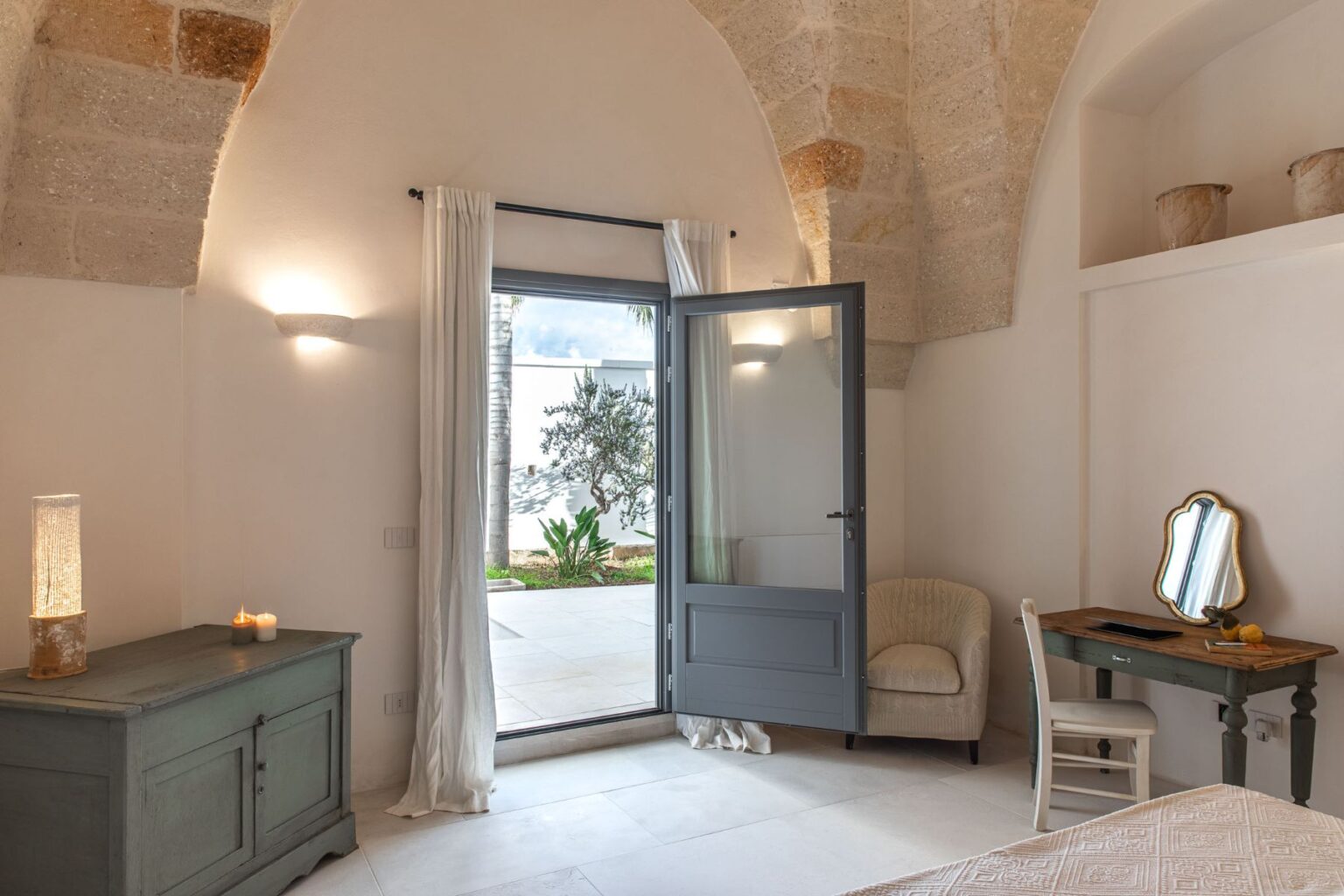 Cozy bedroom with a gray door opening to a patio, featuring a light-colored armchair, a wooden desk with a mirror, and a decorative table with candles. The room showcases natural stone arches and soft lighting, creating a serene atmosphere. | Only on SopranoVillas