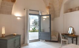 Cozy bedroom with a gray door opening to a patio, featuring a light-colored armchair, a wooden desk with a mirror, and a decorative table with candles. The room showcases natural stone arches and soft lighting, creating a serene atmosphere.