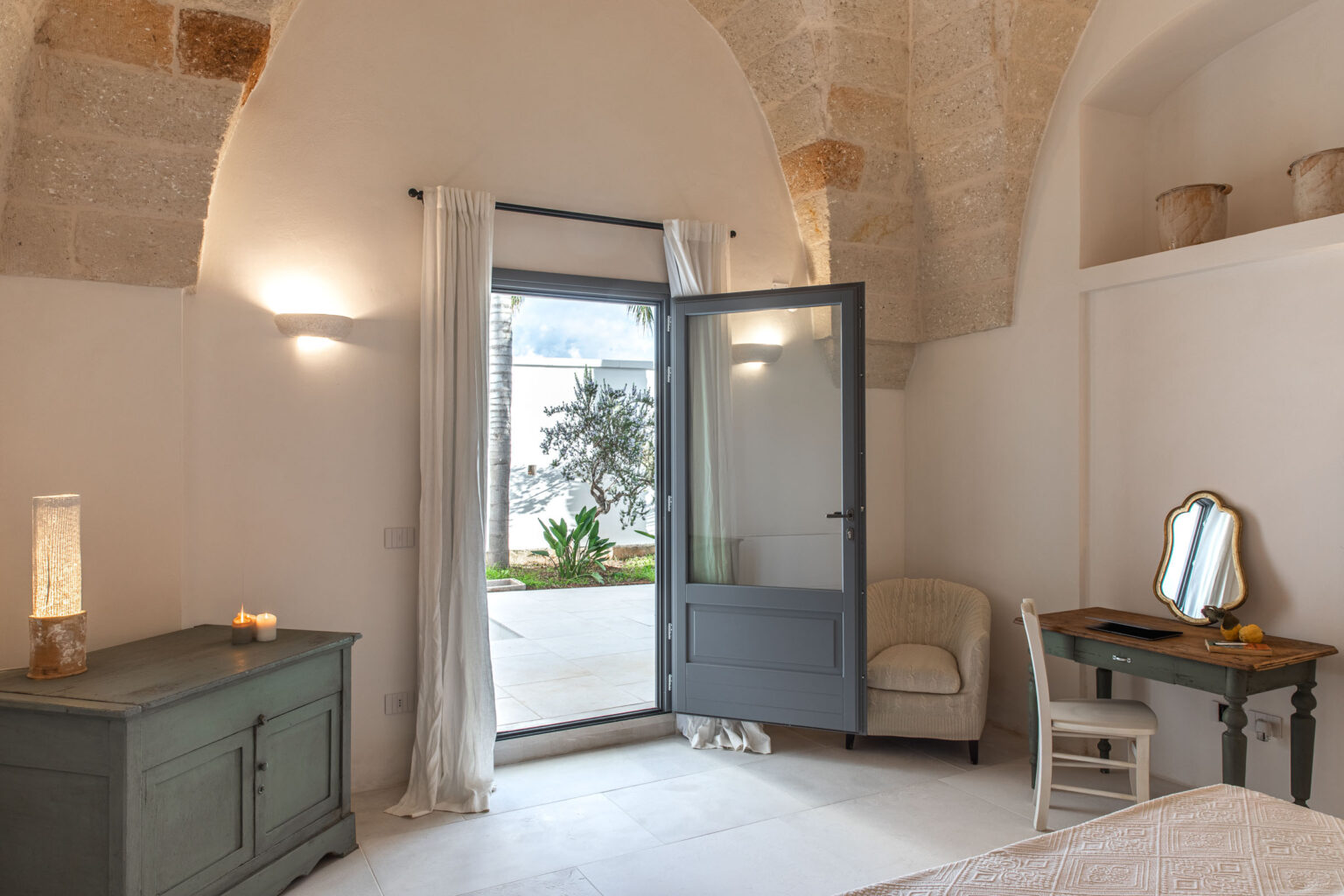 Bright and airy interior of a modern bedroom featuring an arched stone ceiling, an open door leading to an outdoor area, a cozy armchair, a wooden desk with a mirror, and decorative lighting. | Only on SopranoVillas