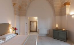Modern bedroom interior featuring a stylish bed with decorative pillows, an elegant wooden chest, and a floral arrangement. The room boasts high stone arch ceilings, soft lighting, and a minimalist design, creating a serene and inviting atmosphere. A glimpse of the adjoining bathroom area can be seen through the doorway.