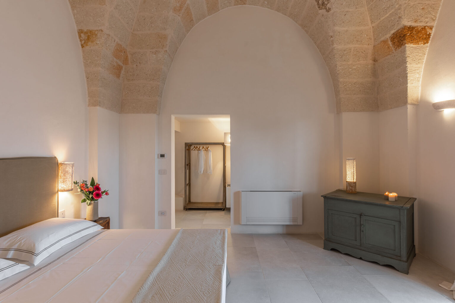 Spacious and elegantly designed bedroom featuring an arched ceiling, neutral color palette, and tasteful decor, with a cozy bed, a stylish dresser, and a doorway leading to a modern bathroom. | Only on SopranoVillas