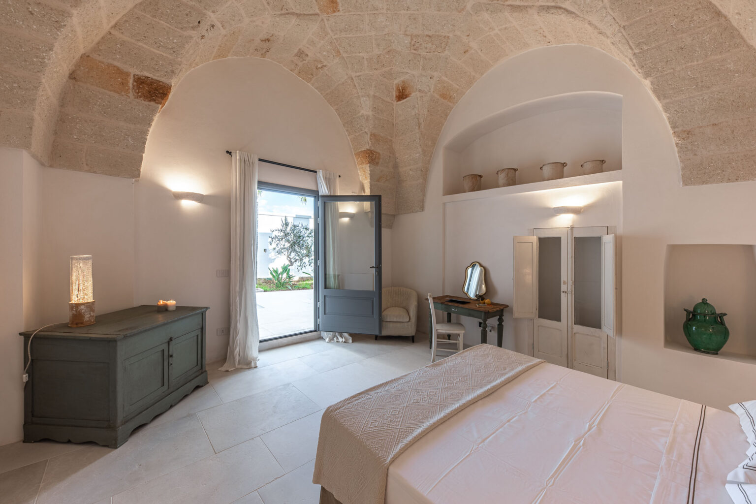 Bright and airy bedroom featuring a vaulted stone ceiling, a cozy bed with a decorative throw, and a vintage green chest. The room includes a small desk with a mirror, a comfortable chair, and an open door leading to a serene outdoor space with greenery. Soft lighting adds to the inviting atmosphere. | Only on SopranoVillas