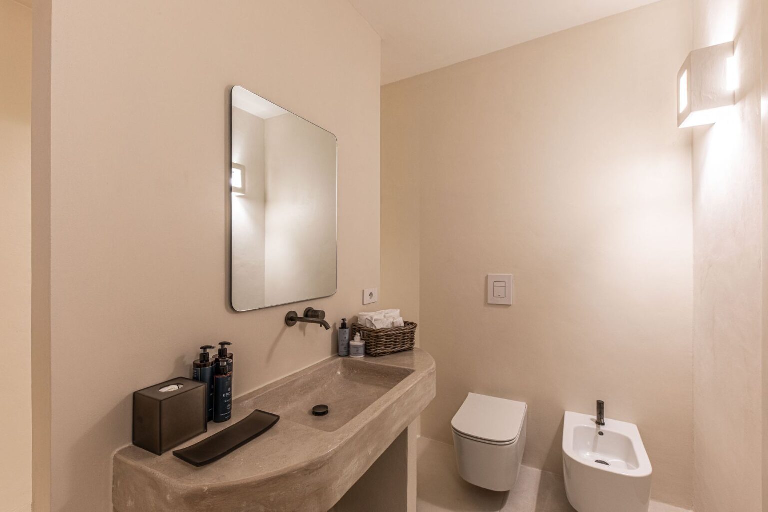 Modern bathroom featuring a minimalist vanity with a stone sink, large mirror, and stylish light fixtures. The space includes a contemporary toilet and bidet, complemented by natural tones and organized toiletries for a clean, serene atmosphere. | Only on SopranoVillas