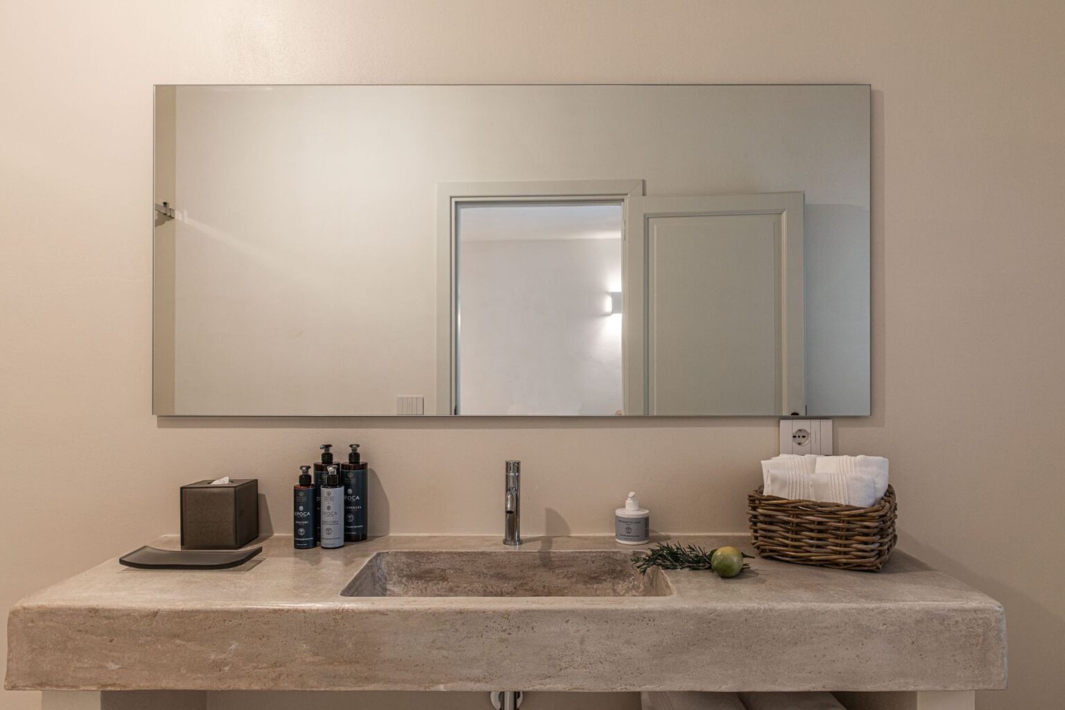 Modern bathroom sink with a minimalist design, featuring a large mirror, decorative toiletries, and organic elements like an apple and rosemary, enhancing the tranquil ambiance. | Only on SopranoVillas