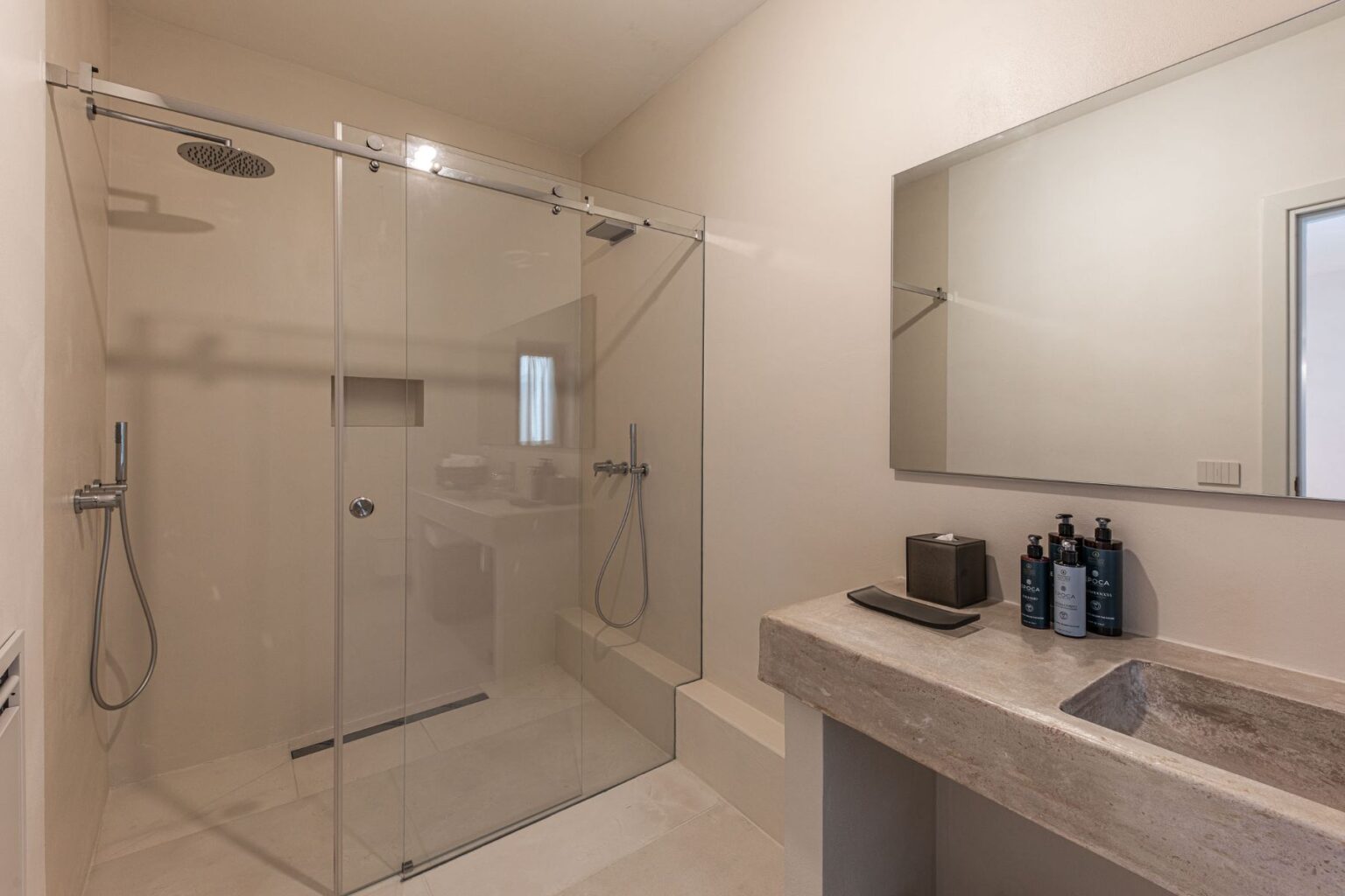 Modern bathroom interior featuring a spacious walk-in shower with transparent glass doors, minimalist design, and a sleek stone sink. The area includes neatly arranged bath products on the counter and a large mirror, creating a clean and contemporary ambiance. | Only on SopranoVillas