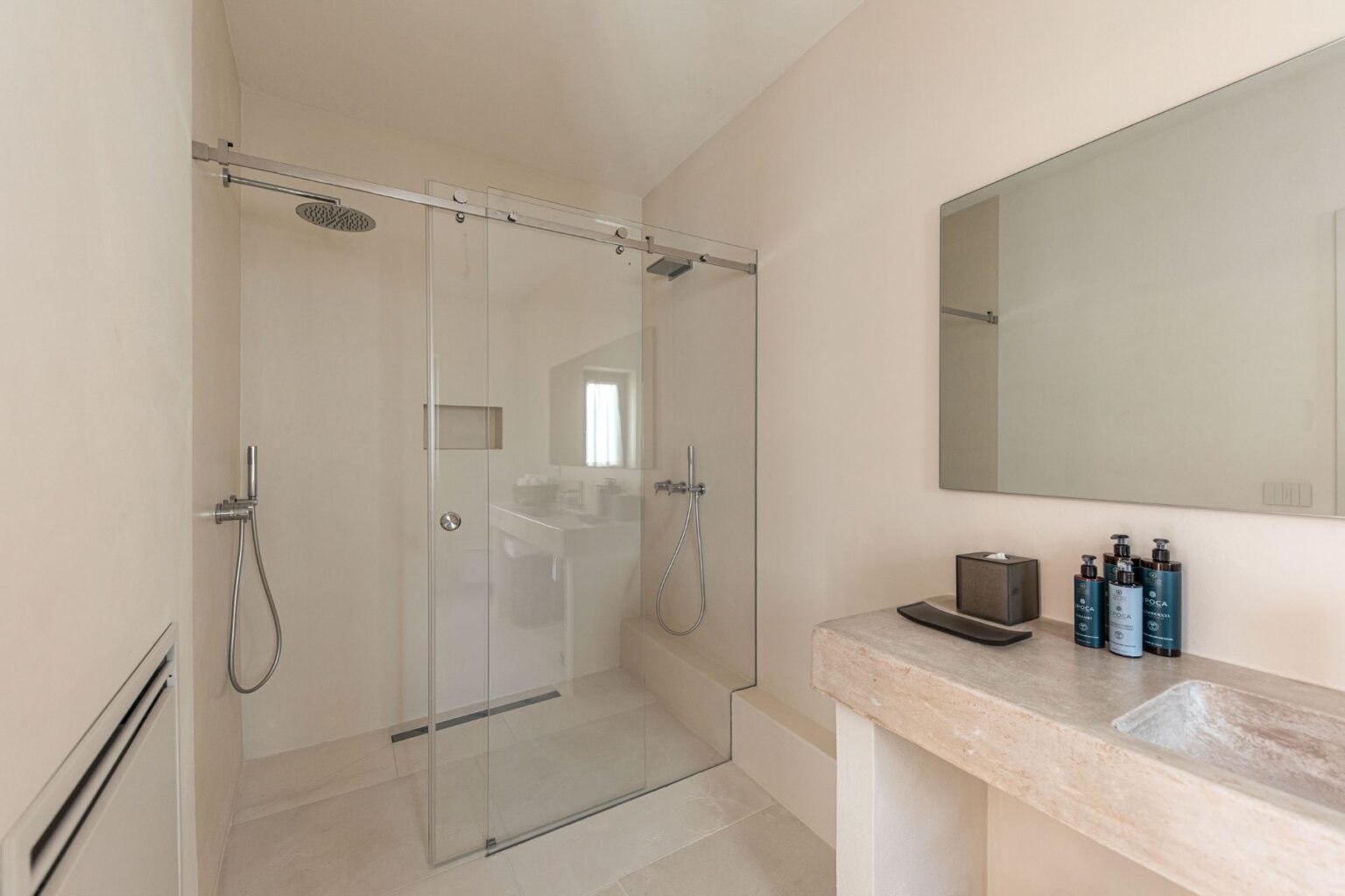 Modern bathroom with a glass shower enclosure, light-colored walls, and a minimalist design featuring a stone countertop with toiletries and a mirror. The space emphasizes cleanliness and contemporary style. | Only on SopranoVillas