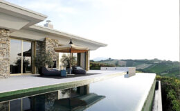 Modern villa with stone accents featuring a spacious outdoor deck, stylish lounge chairs, and an umbrella beside a reflective infinity pool, overlooking lush vineyards and rolling hills.