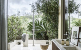 Modern bathroom with a large window overlooking a lush garden, featuring a freestanding bathtub, wooden vanity, and an outdoor shower. The design emphasizes natural light and greenery, creating a serene atmosphere.