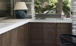 Modern outdoor kitchen featuring a sleek stainless steel grill, dark wooden cabinetry, and a stylish lamp. Surrounded by tropical plants, the space offers a serene atmosphere for dining and entertaining.