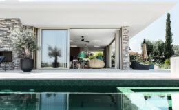 Modern villa with a pool, featuring large glass doors that open to an elegantly designed interior with stone accents and greenery. The outdoor space includes stylish furniture and lush landscaping, creating a serene and inviting atmosphere.