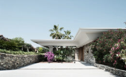 Modern architectural design featuring a stone-clad house surrounded by lush tropical landscaping, including palm trees and vibrant flowering plants, under a clear blue sky.