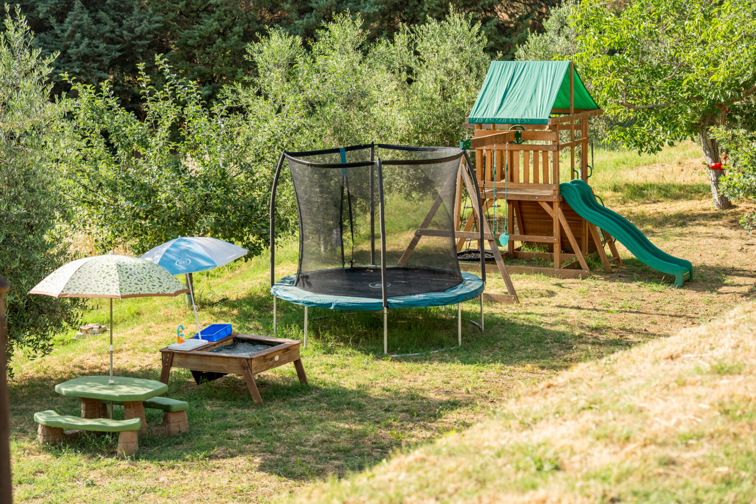 Children's outdoor play area featuring a trampoline, sandbox with toys, picnic table, and a wooden play structure with a slide and swings, surrounded by lush greenery and sunny skies. | Only on SopranoVillas