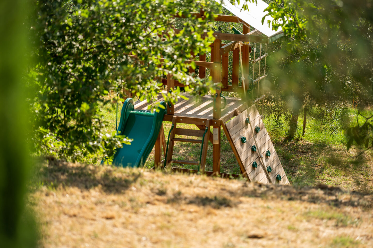 Playground structure with a green slide, climbing wall, and wooden frame, surrounded by trees and grass in a sunny outdoor setting. Ideal for children's play and outdoor activities. | Only on SopranoVillas