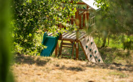 Playground structure with a green slide, climbing wall, and wooden frame, surrounded by trees and grass in a sunny outdoor setting. Ideal for children's play and outdoor activities.