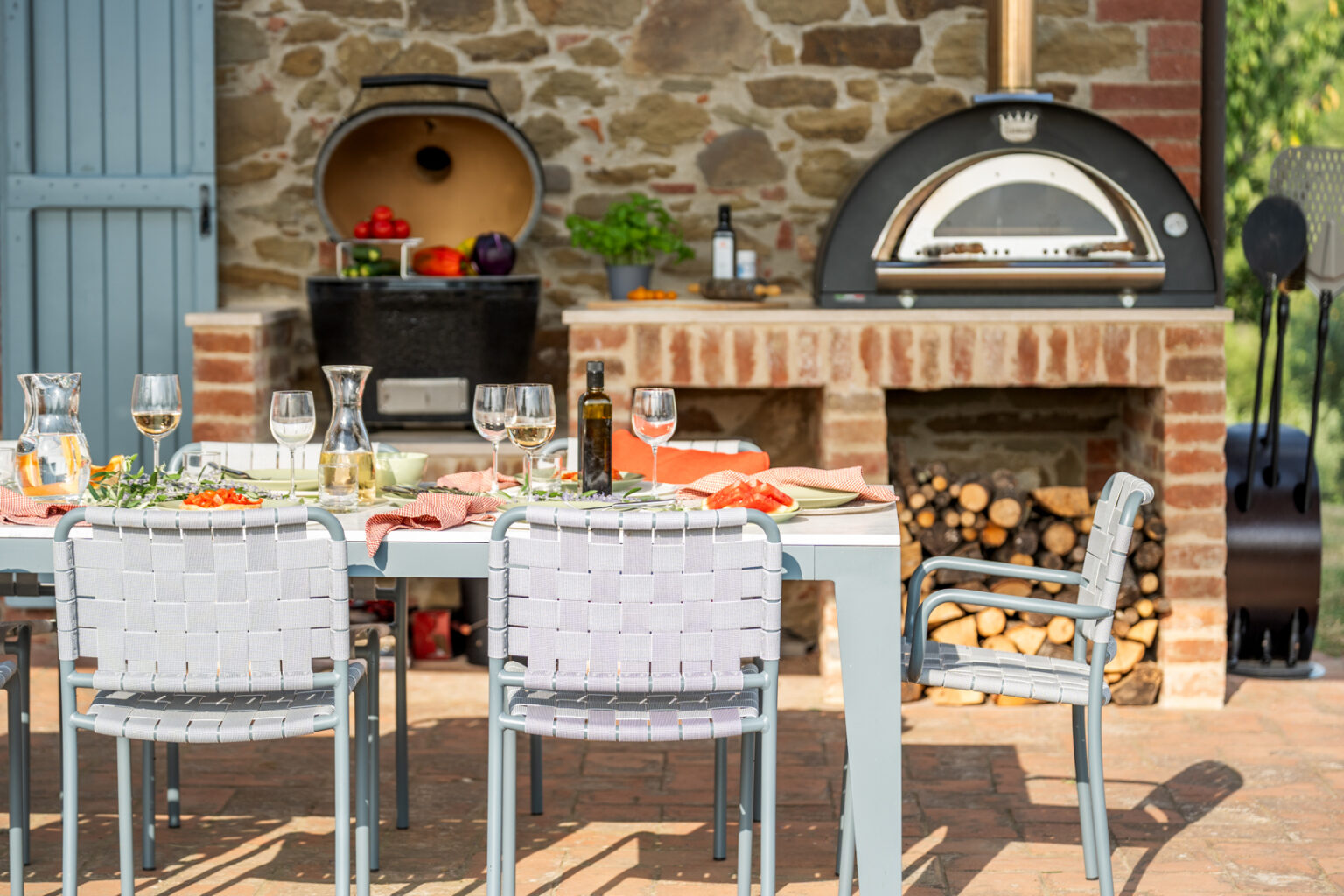 Outdoor dining setup featuring a modern table with woven chairs, elegantly set with wine glasses, a carafe, and a spread of fresh food. An outdoor kitchen with a pizza oven and a storage area for firewood is visible in the background, complementing the rustic stone wall and blue door. Ideal for al fresco gatherings and culinary experiences. | Only on SopranoVillas