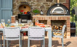 Outdoor dining setup featuring a modern table with woven chairs, elegantly set with wine glasses, a carafe, and a spread of fresh food. An outdoor kitchen with a pizza oven and a storage area for firewood is visible in the background, complementing the rustic stone wall and blue door. Ideal for al fresco gatherings and culinary experiences.