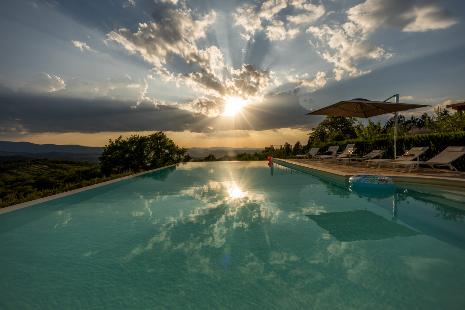 Stunning sunset over an infinity pool with reflective water, surrounded by lush greenery and lounge chairs, creating a serene and picturesque atmosphere in a scenic landscape. | Only on SopranoVillas