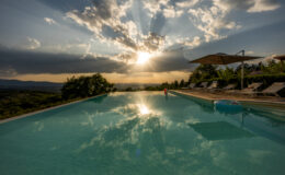 Stunning sunset over an infinity pool with reflective water, surrounded by lush greenery and lounge chairs, creating a serene and picturesque atmosphere in a scenic landscape.