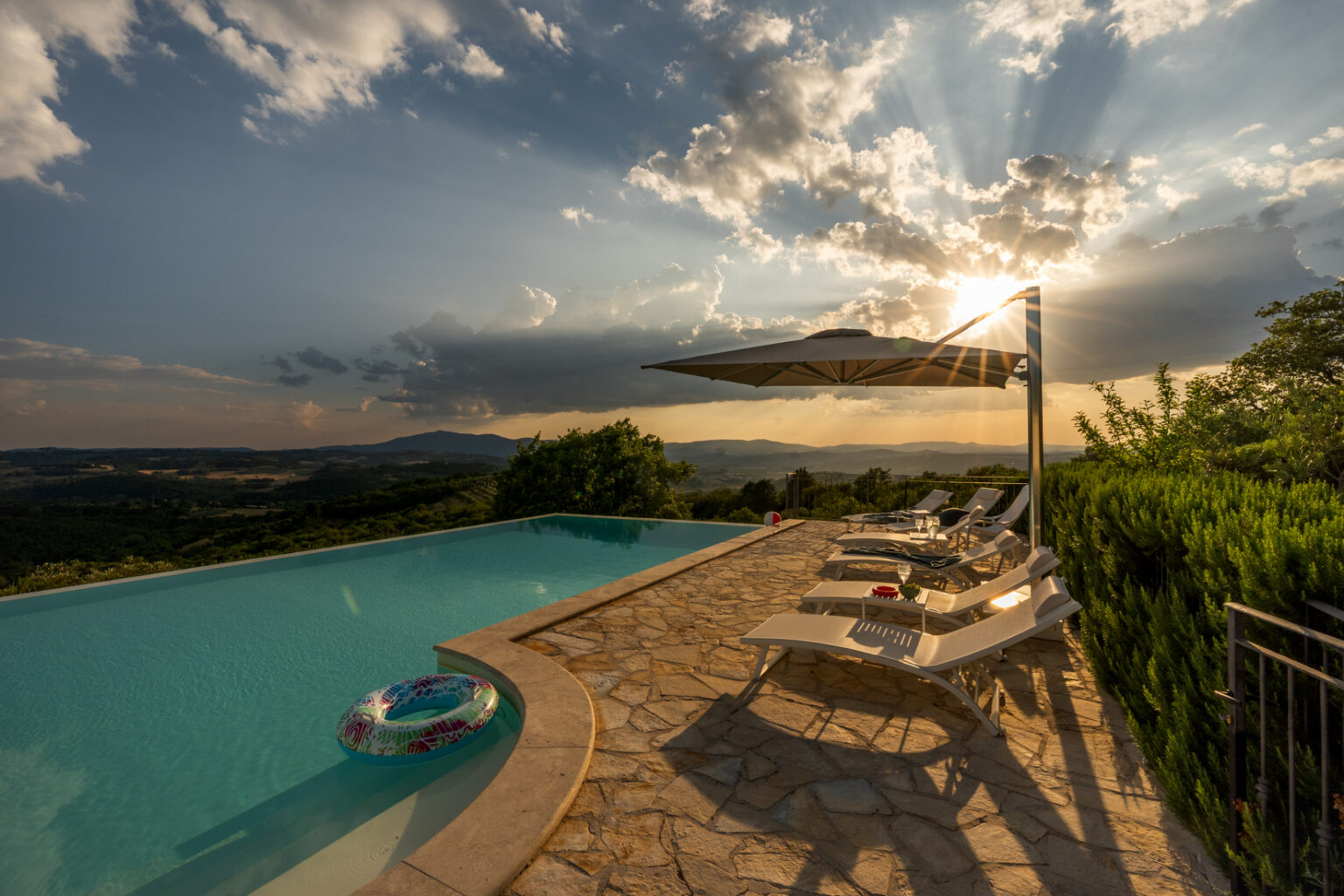Stunning sunset view over a tranquil infinity pool surrounded by lush hills, featuring sun loungers and an umbrella, ideal for relaxation. The vibrant sky with dramatic clouds enhances the serene atmosphere of this luxury outdoor space. | Only on SopranoVillas