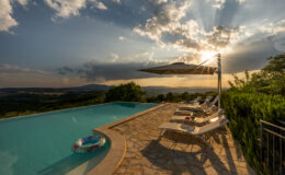 Stunning sunset view over a tranquil infinity pool surrounded by lush hills, featuring sun loungers and an umbrella, ideal for relaxation. The vibrant sky with dramatic clouds enhances the serene atmosphere of this luxury outdoor space.