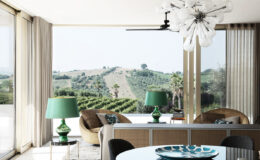 Modern living room with large windows showcasing a scenic view of rolling hills and greenery. The interior features stylish furnishings, including a cozy brown chair, a round dining table, and vibrant green lamps. Natural light floods the space, enhancing the contemporary design and serene atmosphere.