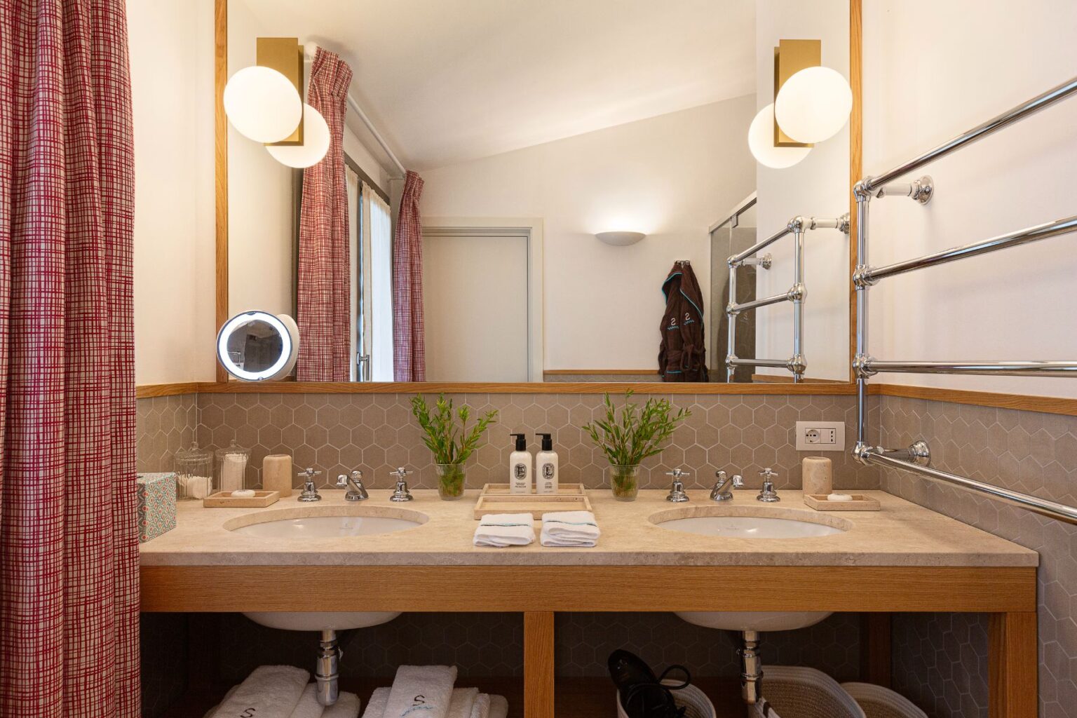 Modern bathroom interior featuring double sinks, stylish lighting, and a warm color palette. The countertop is adorned with potted plants, toiletries, and neatly arranged towels. Decorative curtains and a mirror enhance the contemporary aesthetic, while a towel rack and robe add functionality. Perfect for a luxurious home or hotel setting. | Only on SopranoVillas