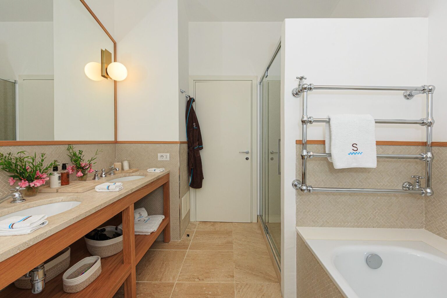 Modern bathroom with dual sinks, stylish lighting, and elegant decor. Features include a glass shower, a bathtub, and organized storage with towels and toiletries. Fresh flowers add a touch of nature to the serene space. | Only on SopranoVillas