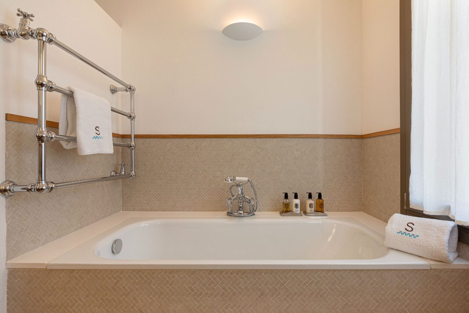Stylish bathroom featuring a modern bathtub, chrome towel rack with a white towel, and neatly arranged toiletries on the edge. Soft natural light filters through a sheer curtain, enhancing the serene ambiance. | Only on SopranoVillas