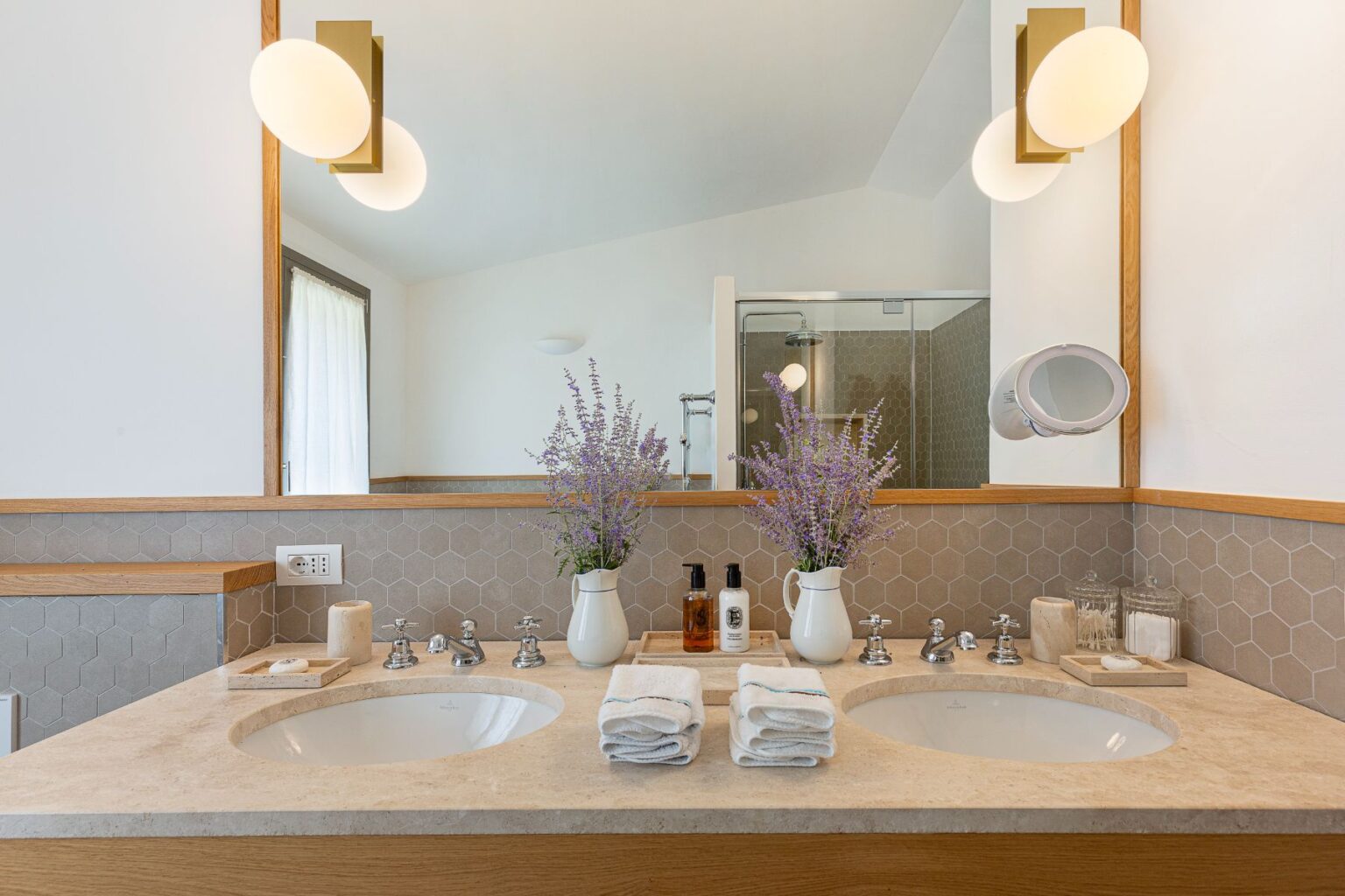 Modern bathroom featuring a double sink vanity with a light-colored stone countertop, stylish hexagonal tile backsplash, and elegant fixtures. Two vases with fresh lavender add a touch of nature, while neatly arranged towels and personal care products enhance the aesthetic. The space is illuminated by contemporary light fixtures and includes a glass shower enclosure in the background. | Only on SopranoVillas