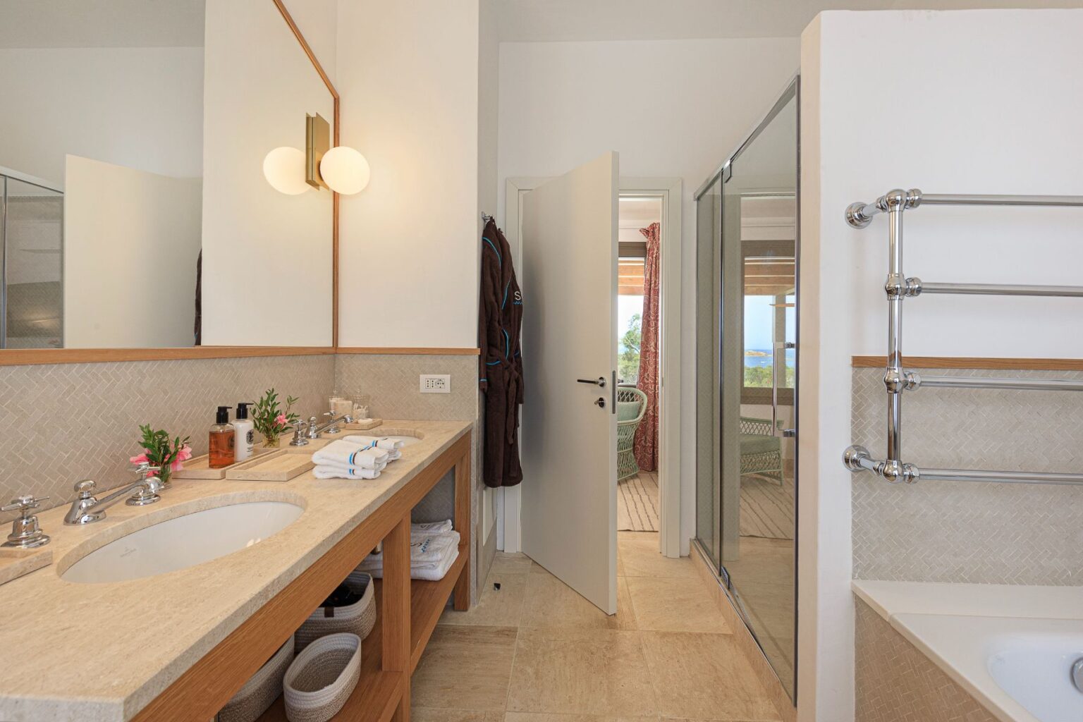Modern bathroom interior featuring a stylish vanity with a round sink, elegant fixtures, and neatly arranged towels. A glass shower enclosure is visible alongside a cozy seating area with a view. Natural light enhances the spacious feel of the room, complemented by decorative plants and warm wood accents. | Only on SopranoVillas