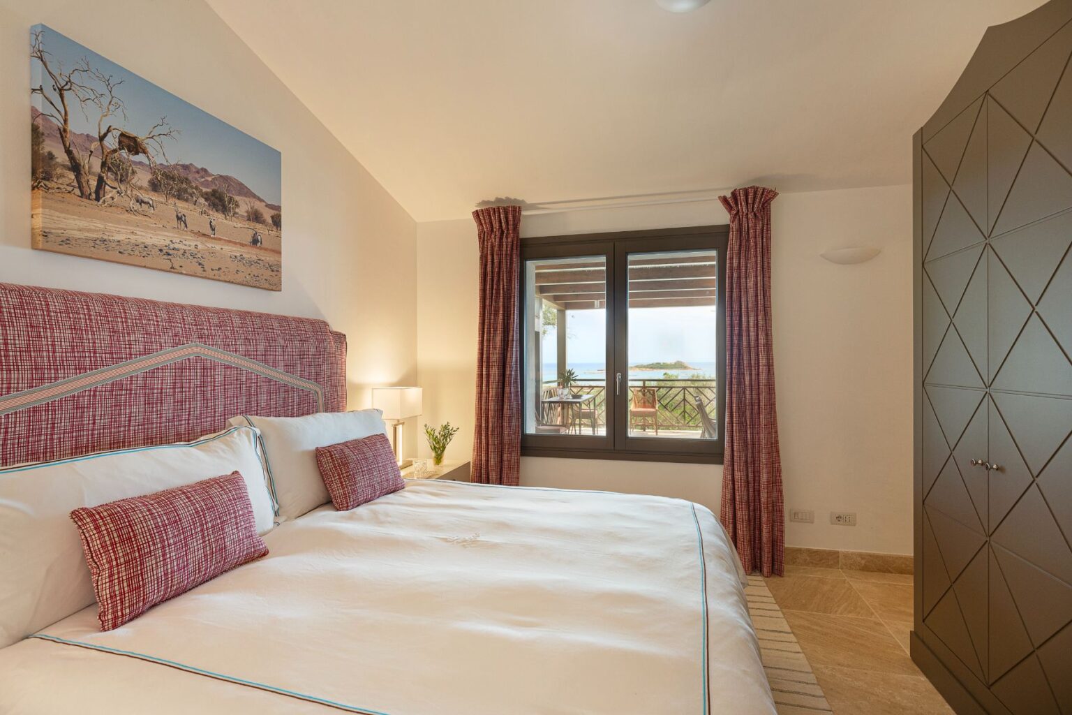 Cozy bedroom featuring a king-sized bed with decorative pillows, a stylish headboard, and a scenic landscape painting. Natural light floods the room through large windows, showcasing a beautiful outdoor view. The space includes elegant curtains, a bedside lamp, and a modern wardrobe, creating a warm and inviting atmosphere. | Only on SopranoVillas