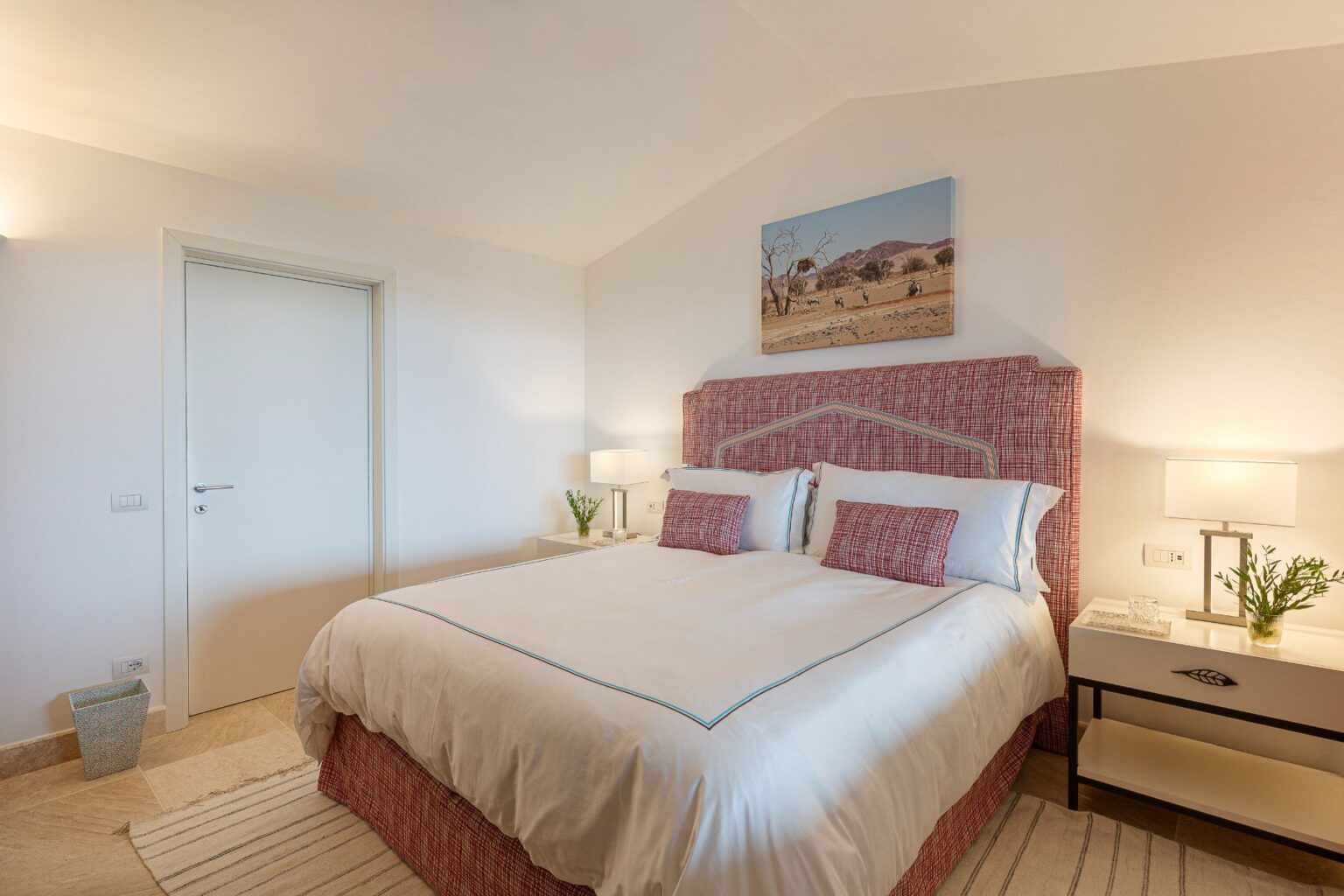 Cozy bedroom featuring a king-sized bed with a patterned headboard, elegant bedding, and decorative pillows. The room includes a bedside table with a lamp and a small plant, along with a framed landscape artwork on the wall. Soft lighting and neutral tones create a relaxing atmosphere. | Only on SopranoVillas