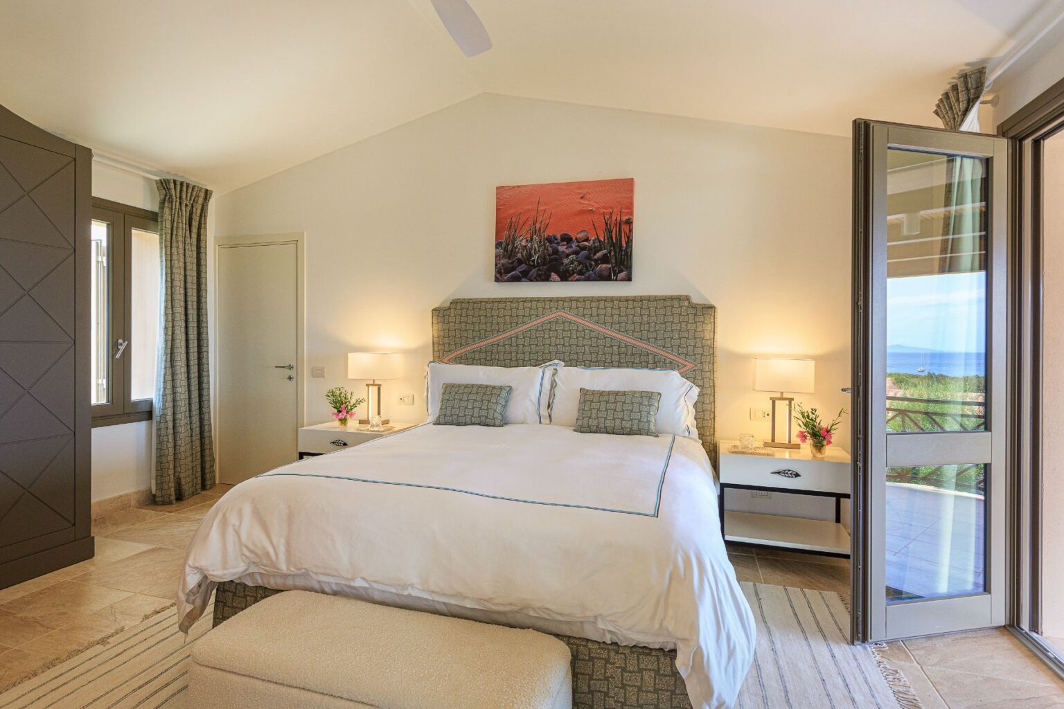 Cozy and modern bedroom featuring a king-sized bed with decorative pillows, bedside lamps, and a scenic view through open balcony doors. The room is adorned with soft curtains and a tasteful piece of artwork above the headboard, complemented by a plush ottoman and a small bouquet of flowers on the nightstand. | Only on SopranoVillas