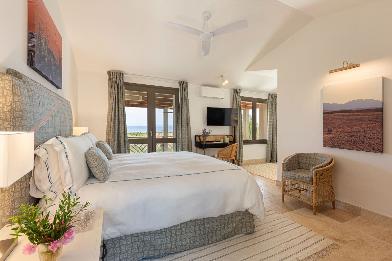 Bright and airy hotel room featuring a king-sized bed, decorative pillows, and a modern design. Large windows provide a scenic view of the outdoors, complemented by elegant curtains. A cozy seating area with rattan chairs and a small table is included, along with a flat-screen TV and air conditioning for comfort. The room is adorned with artwork and a small bouquet of flowers, enhancing the inviting atmosphere. | Only on SopranoVillas