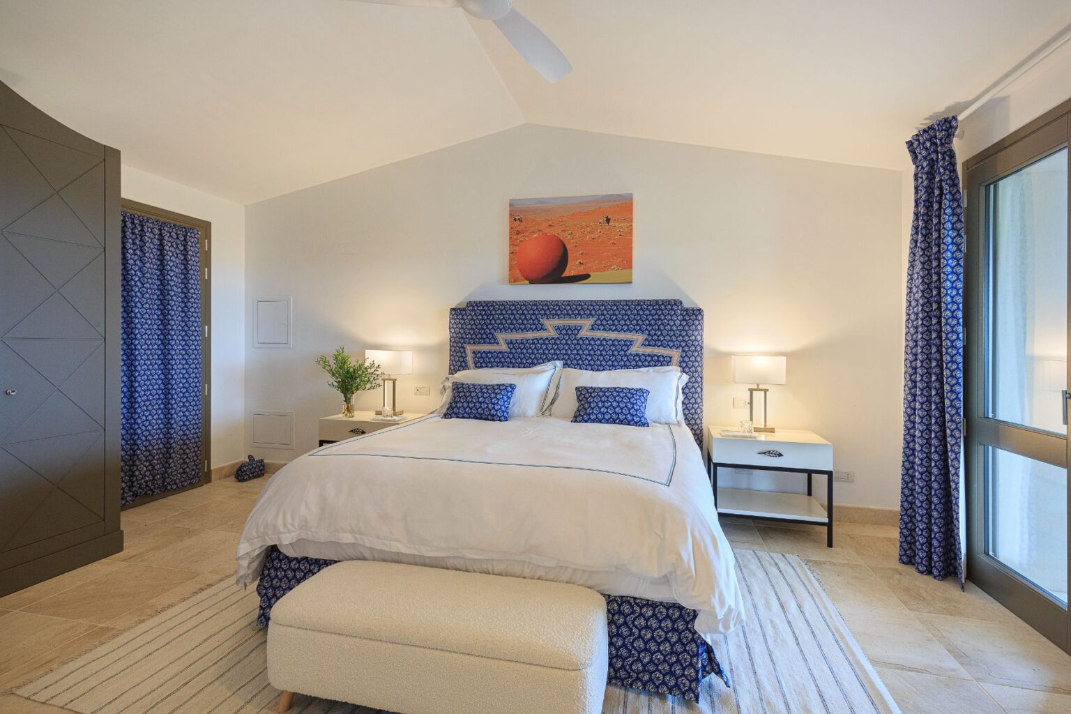 Modern bedroom featuring a plush king-size bed with a blue patterned headboard, decorative pillows, and a cozy bench. The room includes stylish bedside tables with lamps, blue curtains, and a vibrant artwork above the bed, creating a serene and inviting atmosphere. | Only on SopranoVillas