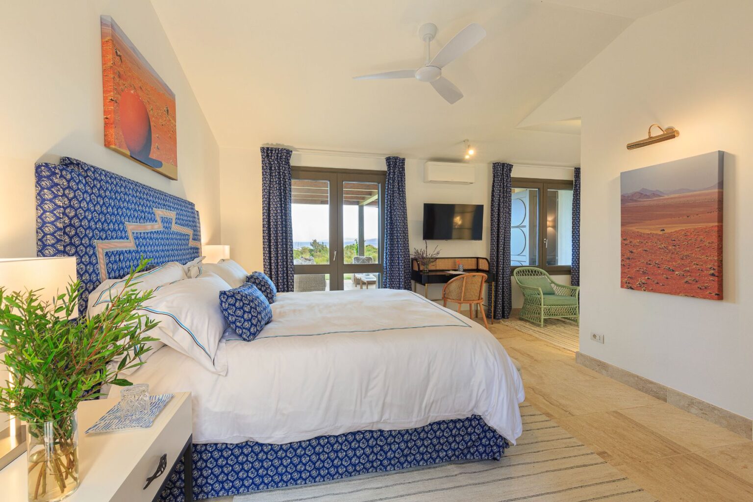 Luxurious hotel room featuring a king-sized bed with blue patterned bedding, stylish bedside lamps, and a decorative plant. The room includes a seating area with a green chair and a flat-screen TV, along with large windows that offer scenic views. Artwork adorns the walls, enhancing the elegant decor. | Only on SopranoVillas