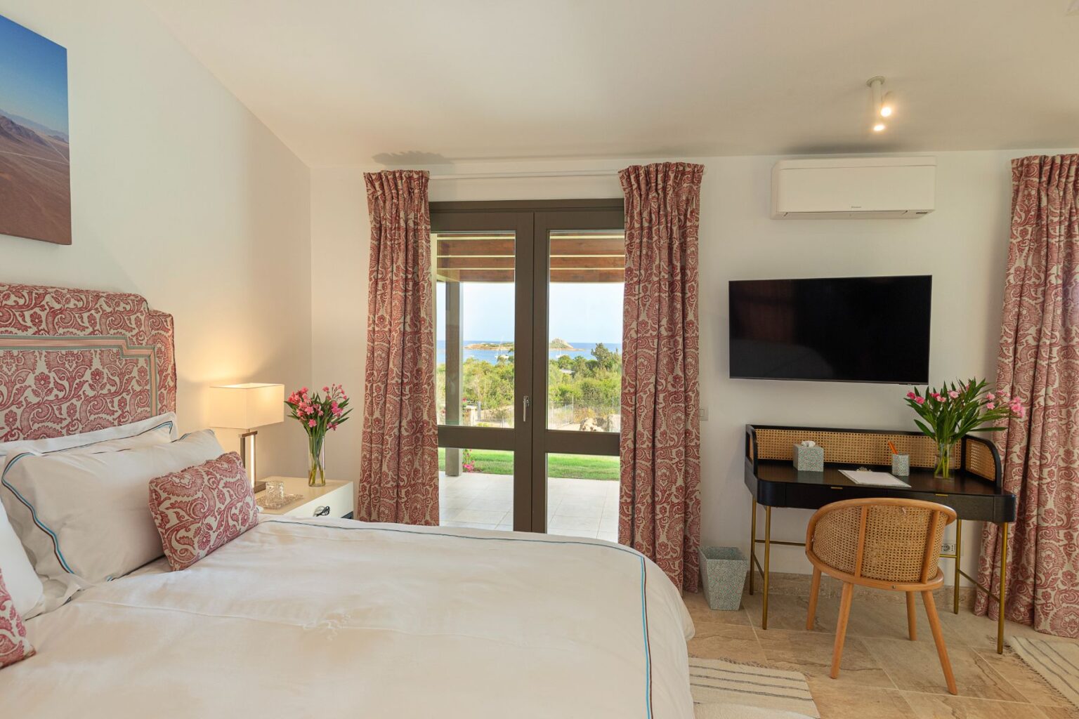 Cozy bedroom featuring a queen-sized bed with decorative pillows, a stylish desk with a chair, and a flat-screen TV. Large windows with patterned curtains offer a view of the outdoors, highlighting lush greenery and a glimpse of the sea. Fresh flowers in a vase add a touch of elegance to the space. | Only on SopranoVillas