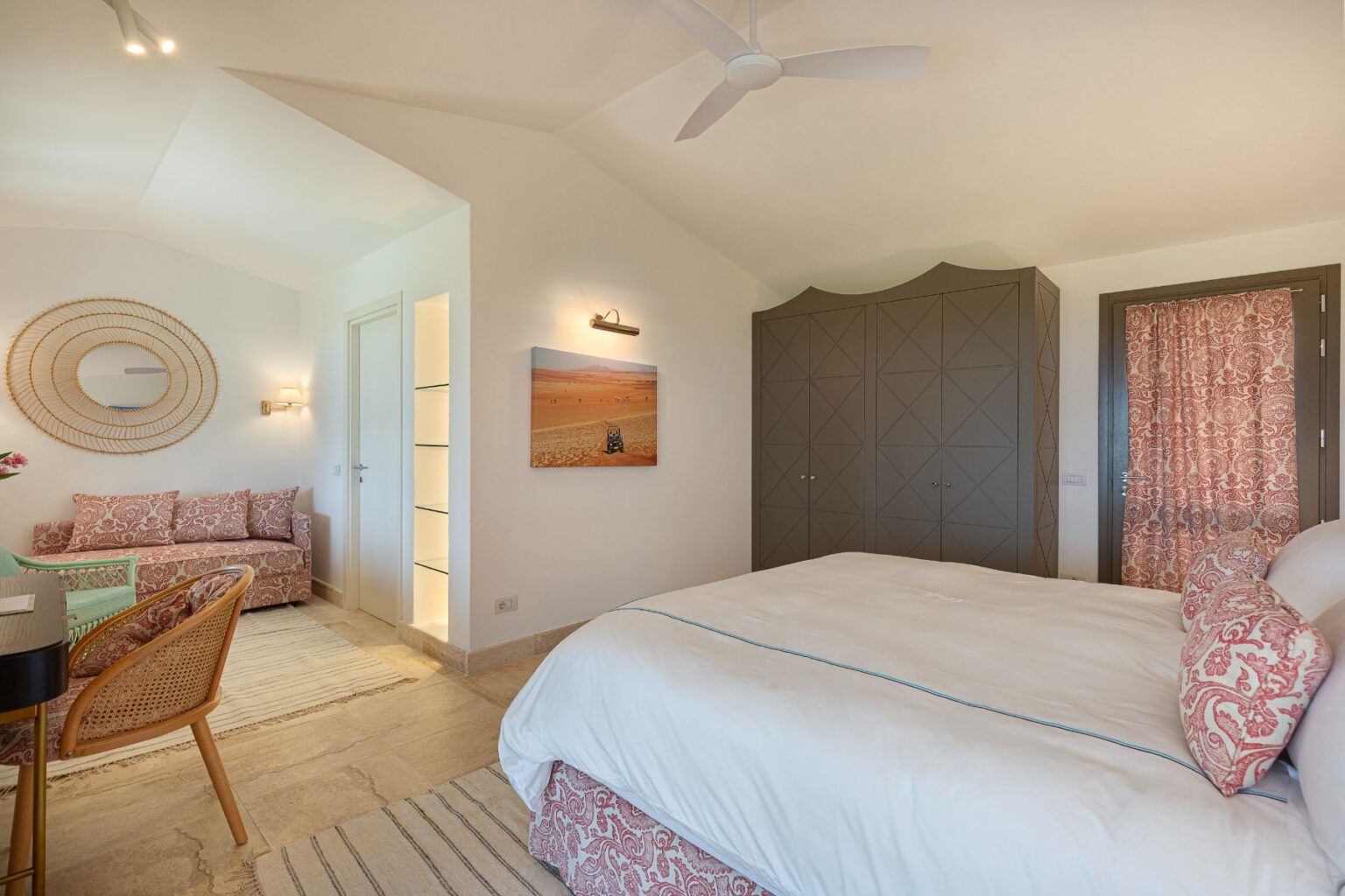 Bright and airy bedroom with a king-sized bed, stylish seating area featuring a patterned sofa, and a modern wardrobe. Decor includes a round mirror and a desert landscape artwork, complemented by natural light and neutral tones. Ideal for relaxation and comfort. | Only on SopranoVillas
